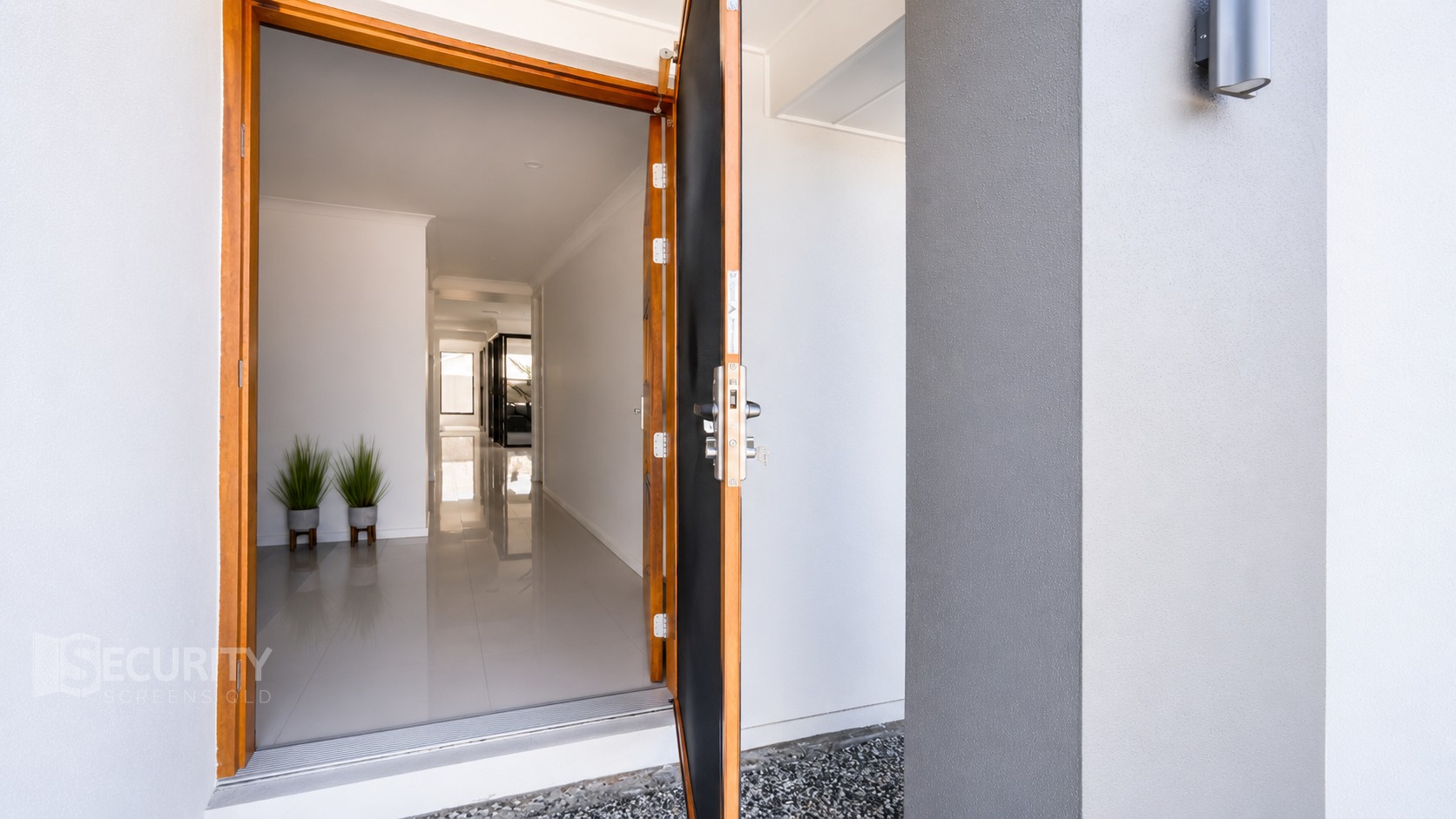 Modern woodgrain framed hinge security door installed at residential front entry in Brisbane, showing stylish dark screen panel with contemporary handle and hallway view.